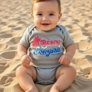 Baby sitting on sand wearing a onesie with beach-themed graphics red white and blue logo